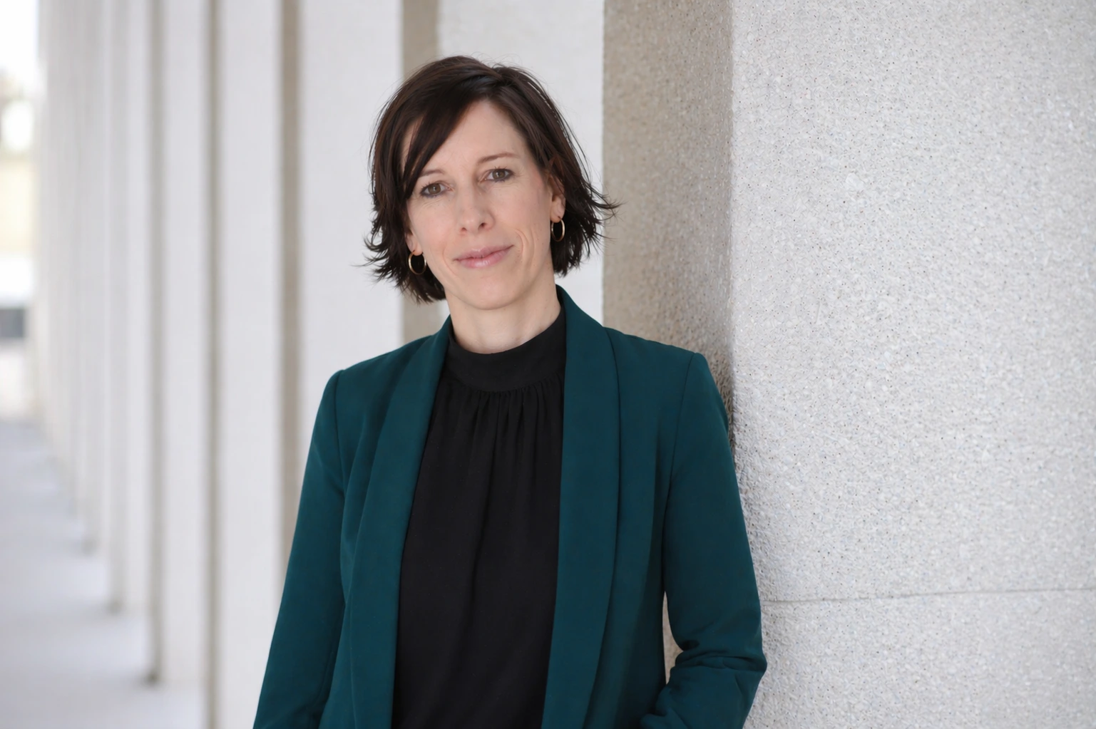 Nina Müller lehnt an einer grauen Säule. Sie trägt ein schwarzes Shirt und einen türkisfarbenen Blazer.
