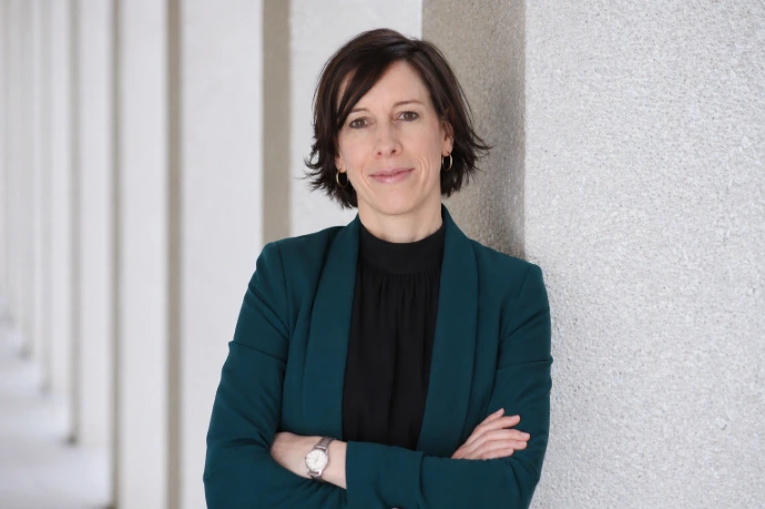 Nina Müller lehnt an einer grauen Säule, die Arme verschränkt. Sie trägt ein schwarzes Shirt und einen türkisfarbenen Blazer.